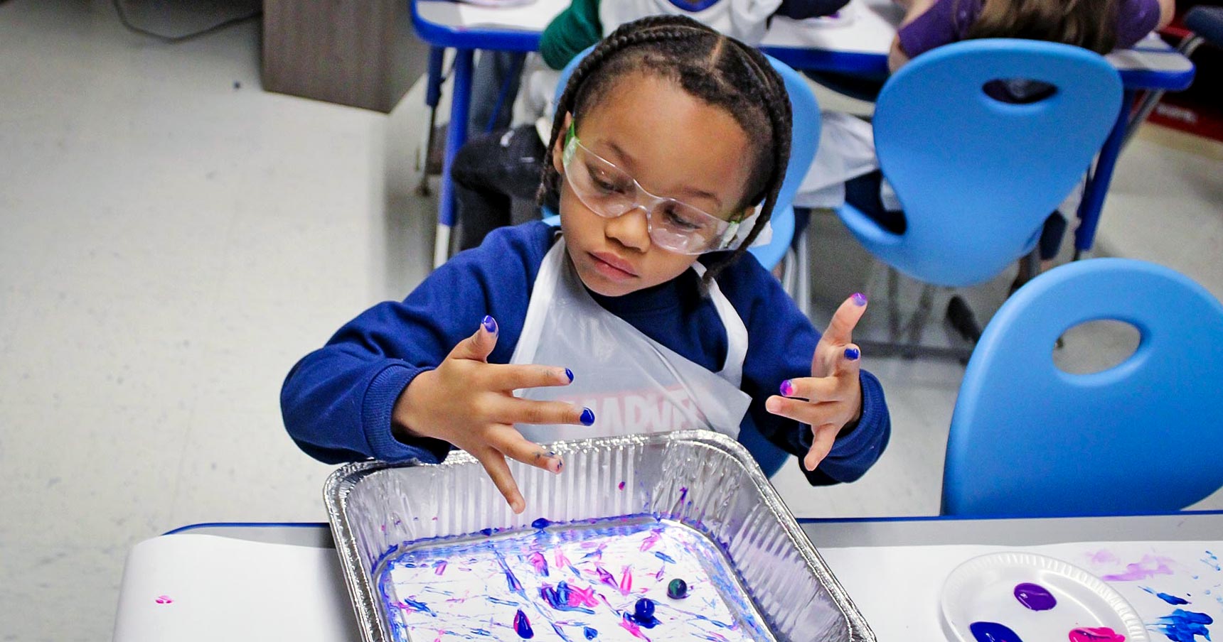 A young boy looks at his fingertips which have purple and pink paint covering them.