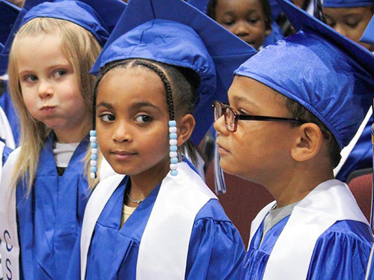 Kids in blue caps and gowns standing side by side as someone speaks at the 2024 Head Start transition into Kindergarten ceremony.
