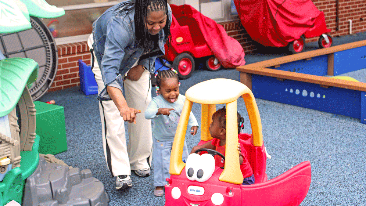 A female Early Head Start Teacher in a denim jacket and white pants points toward a young child in a red shirt sitting inside a red and yellow toy car. Next to the car, a young girl in a light blue shirt smiles and points along with the adult on an outdoor playground with blue safety surfacing.