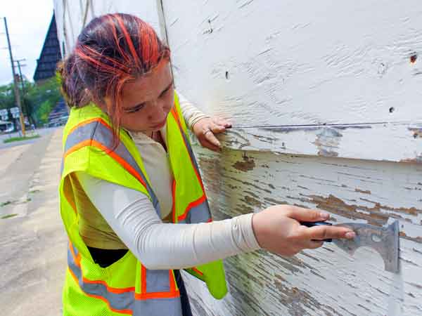 Young woman in yellow safety vest scraping paint off of a building.